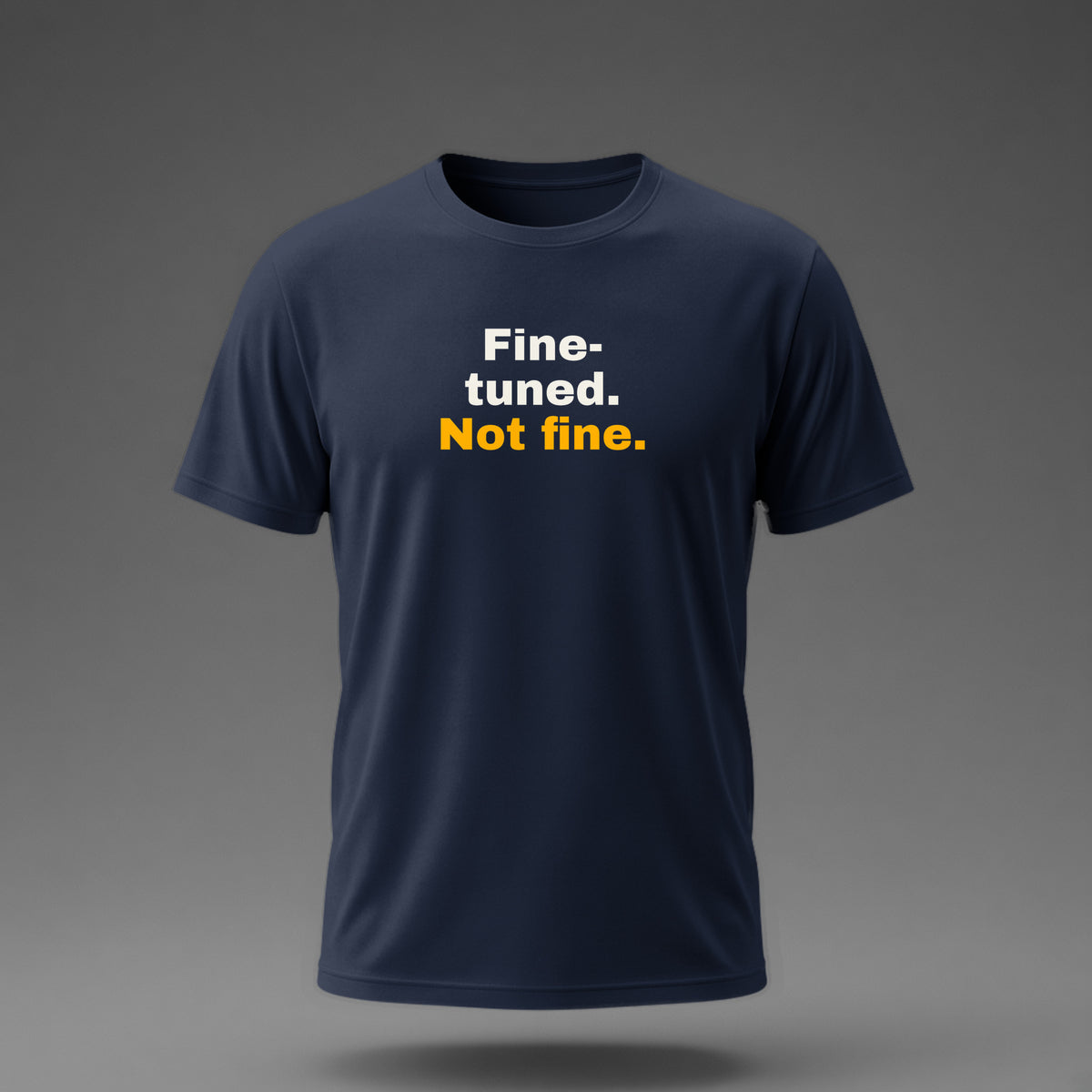 Front view: Navy t-shirt floating on a grey background with 'Fine-tuned. Not fine.' in three heavy lines printed across the chest in off-white and amber.