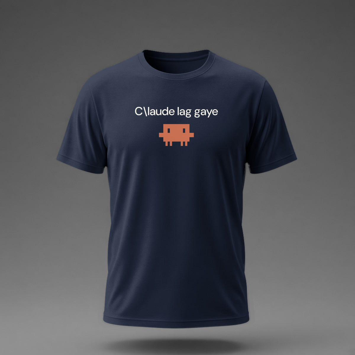 Front view: Navy t-shirt floating on a grey background with a single short lowercase line of off-white text across the chest and a small coral pixel-art critter below it.